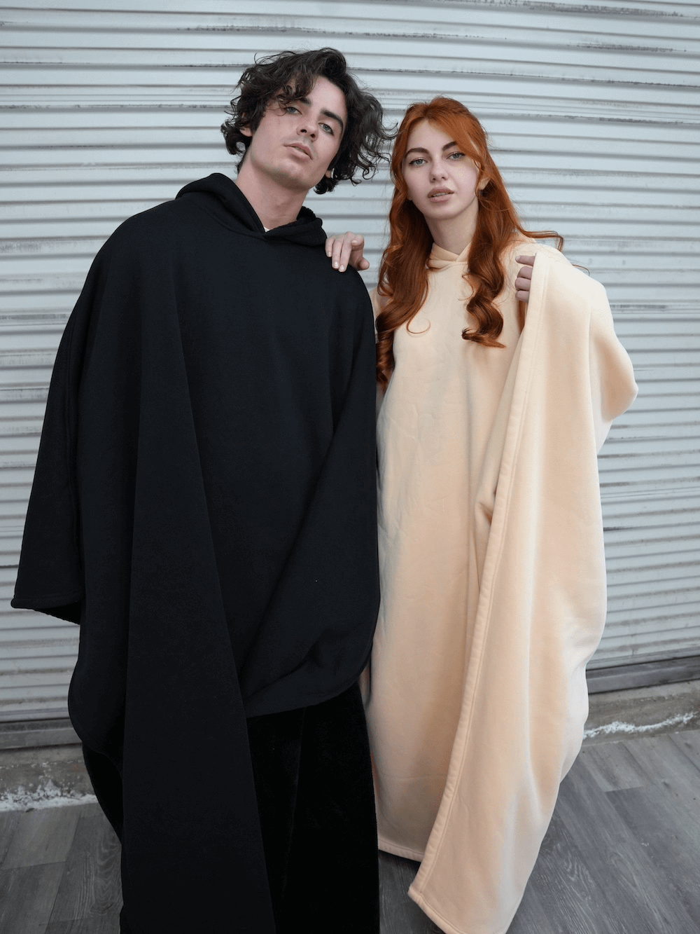 A man in a black hoodie blanket and a woman in a beige hoodie blanket, showcasing comfort and relaxation with the Eden Comfort Hoodie Blanket.